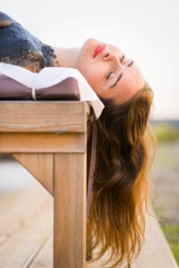 Portrait of a young woman, covered in black health and beauty treatment mud, eyes closed, lying on her back on a massage table, enjoying the experience of the therapy and being outdoors at sunset.