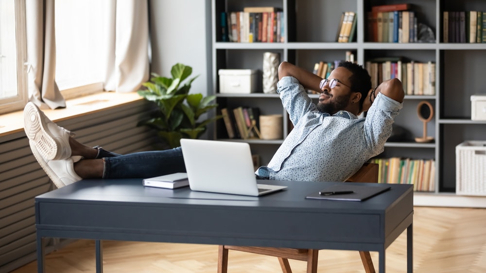 A young man working from home at his desk takes a break with his feet on the desk and his arms behind his head